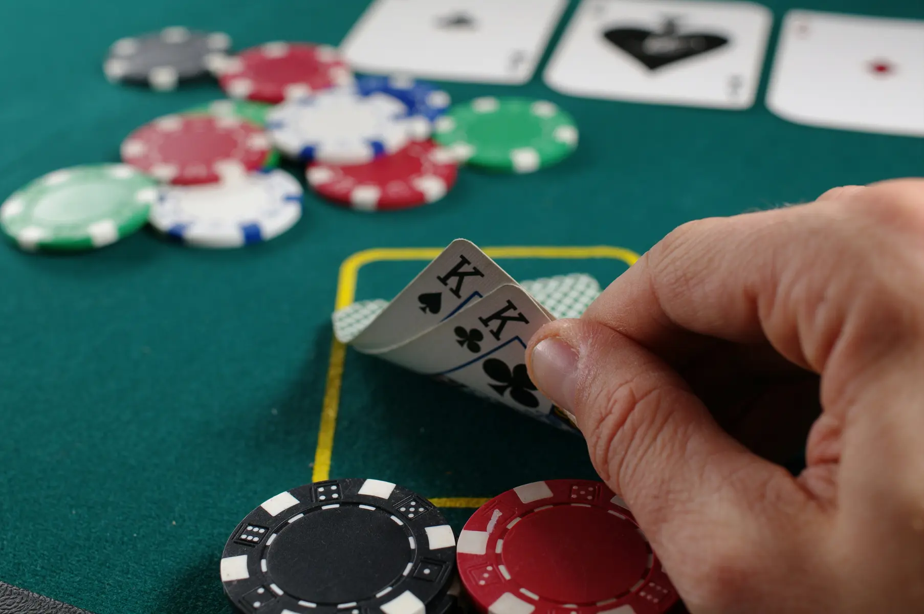 Een hand die twee schoppen koningen vasthoudt op een groene pokertafel met fiches en kaarten.